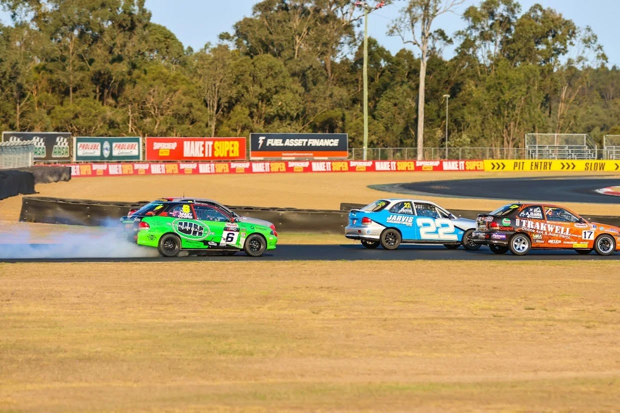 Race cars on a track during a race with trees and sponsor signs in the background.