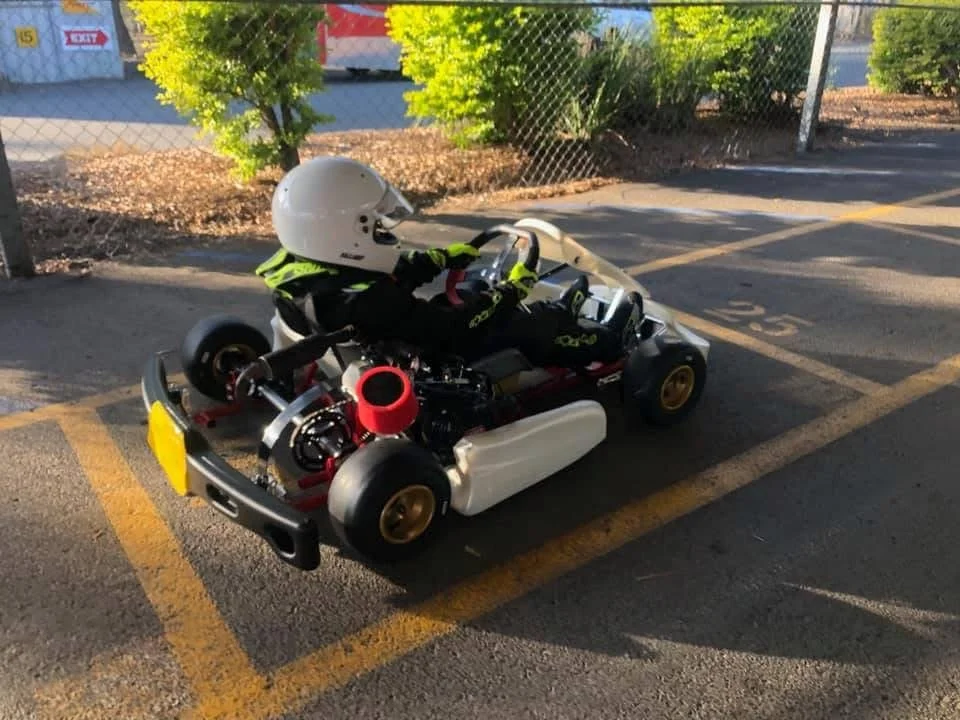 Small go-kart with a racing helmet inside, parked in a parking lot next to a chain-link fence and trees.