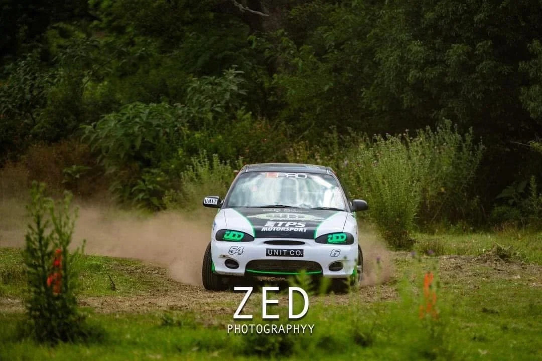 Rally car racing on dirt track surrounded by green trees and bushes, kicking up dust.