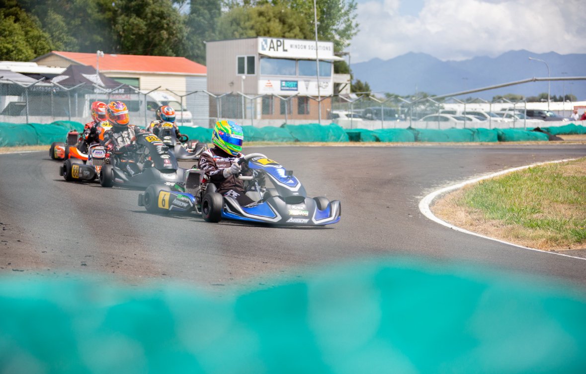 Go-kart racers on an outdoor track, with a group of drivers navigating a turn, fencing, a building in the background, and mountains under a partly cloudy sky.