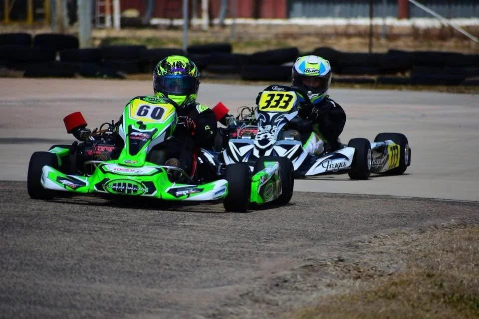 Two go-kart racers wearing helmets and racing suits on a race track, one in front with number 60, the other behind with number 333.