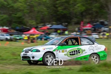 Rally car speeding on a grassy field with spectators and tents in the background