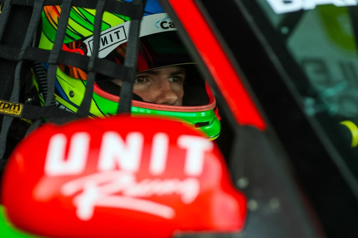 Close-up of a race car driver wearing a vibrant green and black helmet with red accents, seated inside a race car with a safety harness and a red 'UNIT