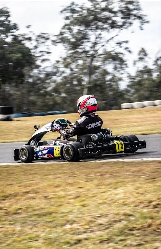 A person racing mini karts on a paved track, wearing a red and white helmet and a black racing suit, with trees and tire barriers in the background.