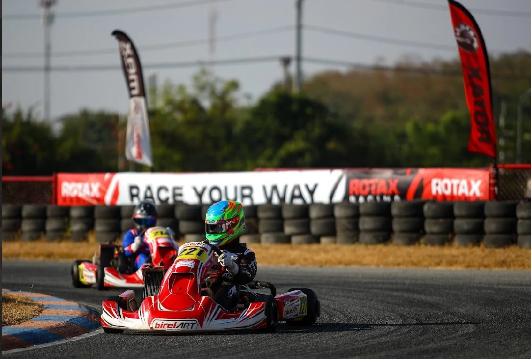 Two go-karts racing on a track around a corner, with a barrier of tires and a banner in the background that reads 'Race Your Way' and has logos for Rotax and Birel ART.
