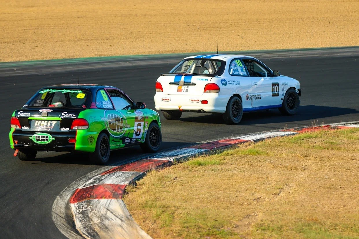 Two race cars on a track, one with green and black livery and the other white with blue stripes, navigating a curve with a red and white curb.