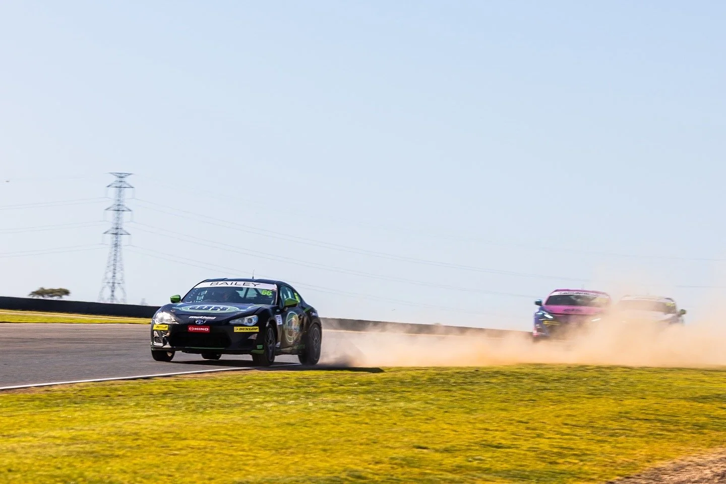 Race cars speeding on a track, kicking up dust, with a clear sky and power lines in the background.