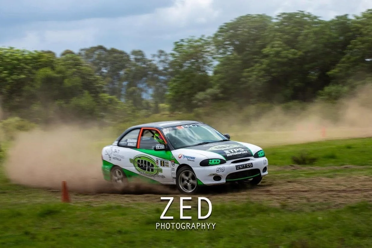 A white race car with green and black accents speeding on a dirt track, kicking up dust, surrounded by green grass and trees.