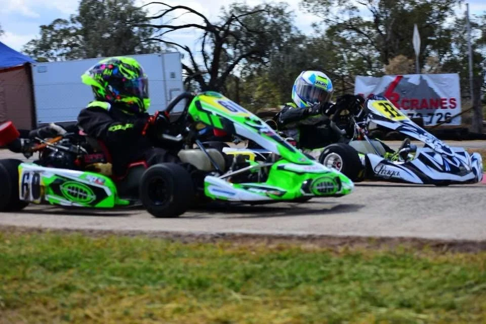 Two go-kart racers wearing helmets and racing suits, competing on an outdoor track.