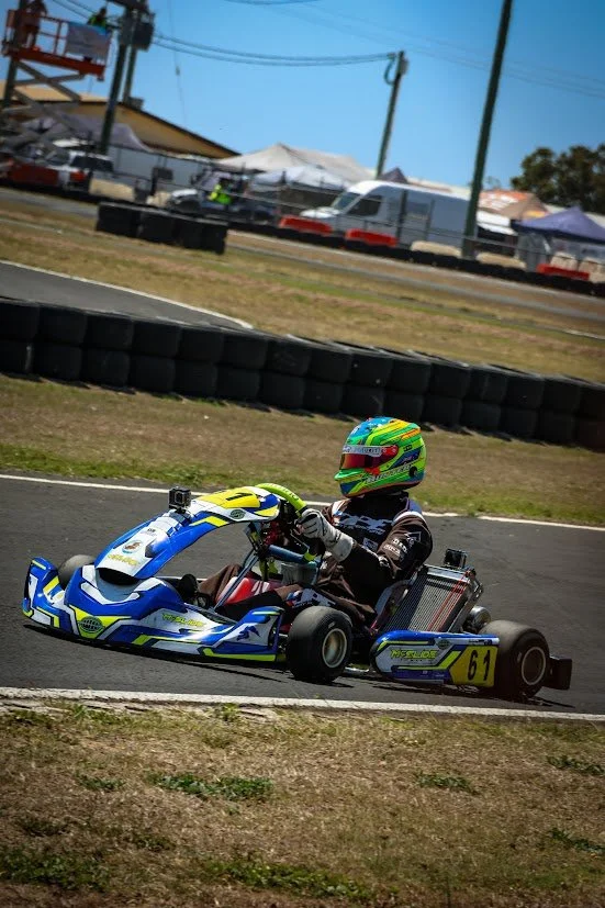 A person racing go-karts on a track, wearing a colorful helmet and black gloves, with tents and vehicles in the background.