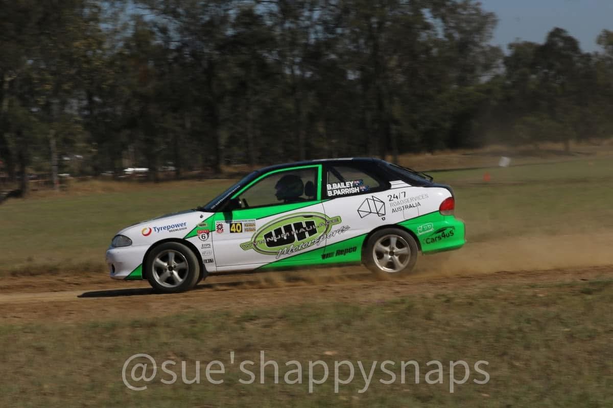 A white and green racing car with sponsor decals speeding on a dirt track, kicking up dust, with trees in the background.