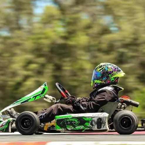 Person in a go-kart wearing a helmet and racing gear, driving on a track with blurred trees in the background.
