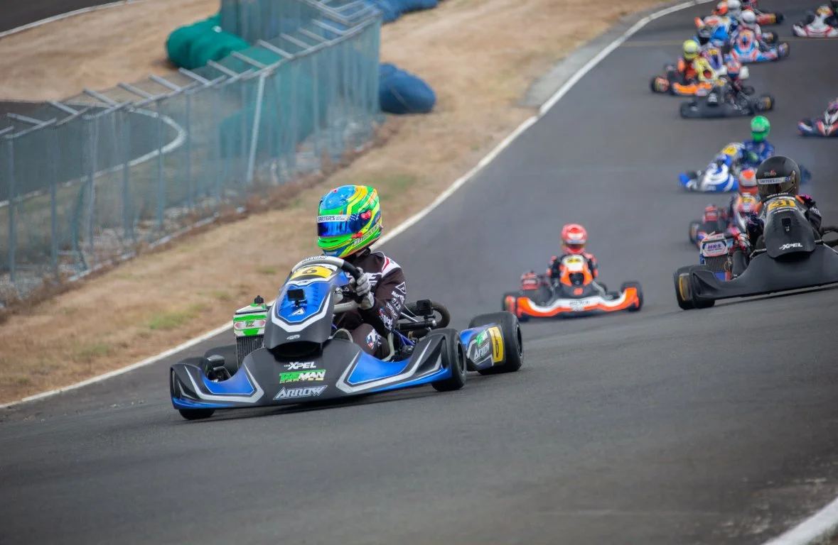 Kart racers driving on a race track with the leading driver in a blue and black kart wearing a colorful helmet.