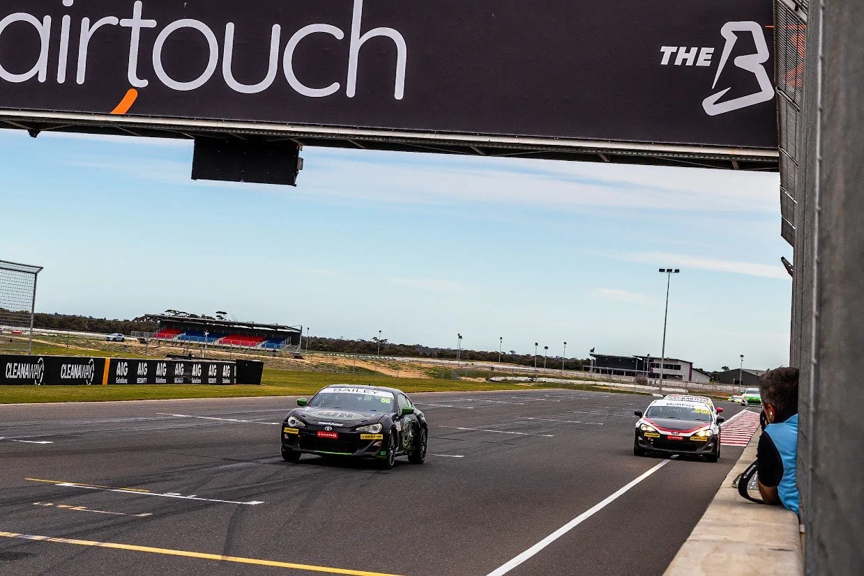 Race cars on a track with sponsor banners, a person in a blue shirt watching from the sidelines, and grandstands in the background.