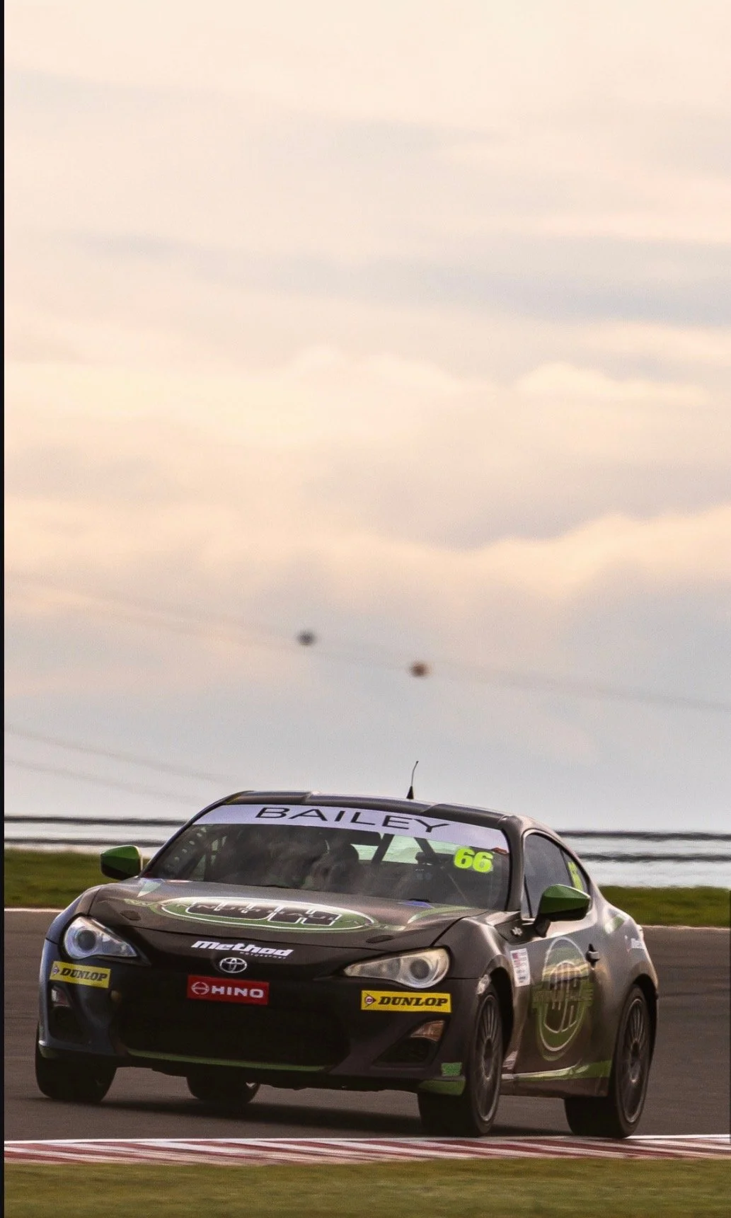 Black race car with the number 66 and sponsor logos, on a racing track with a cloudy sky