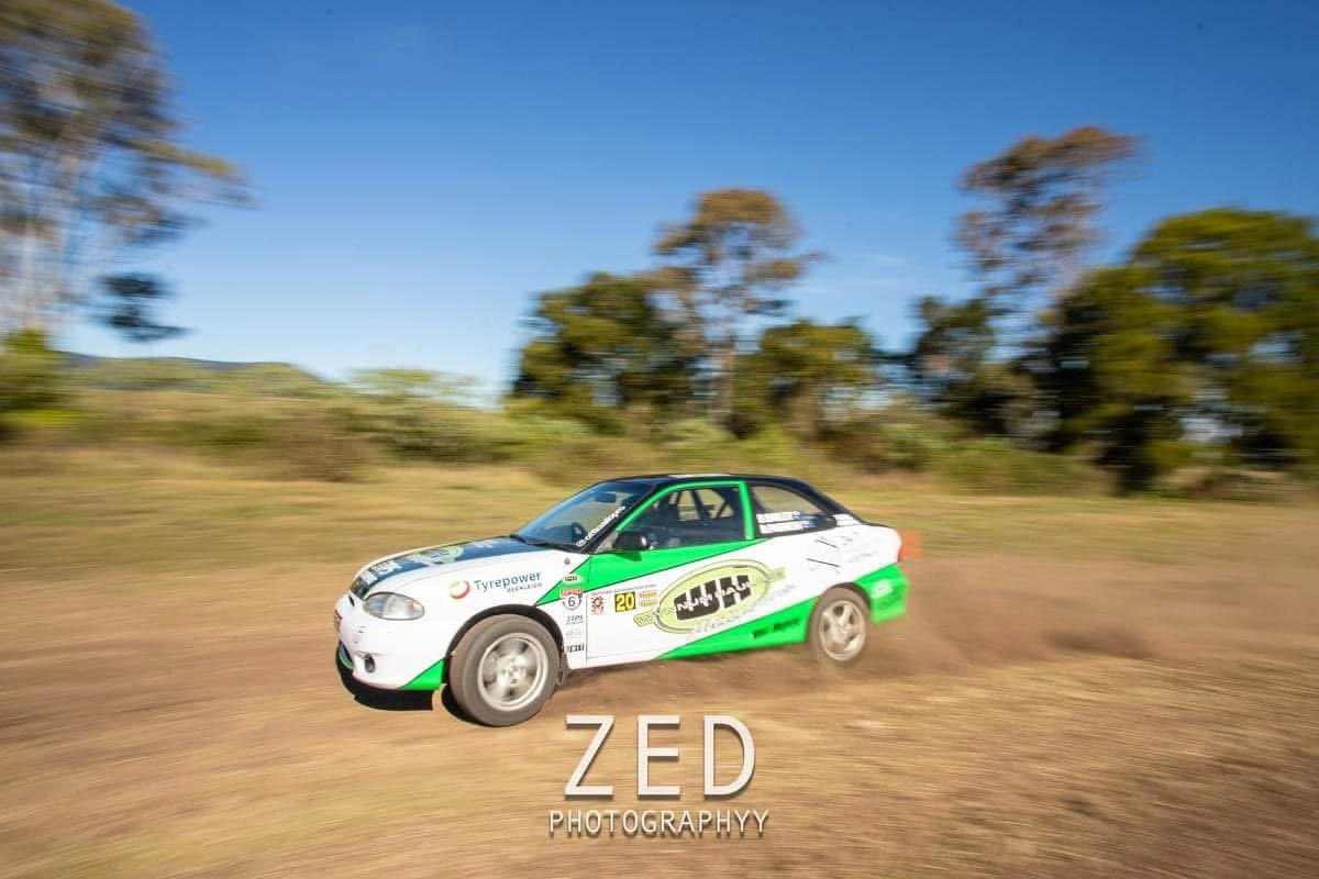 A white rally car with green accents speeds along a dirt road with trees and a blue sky in the background.