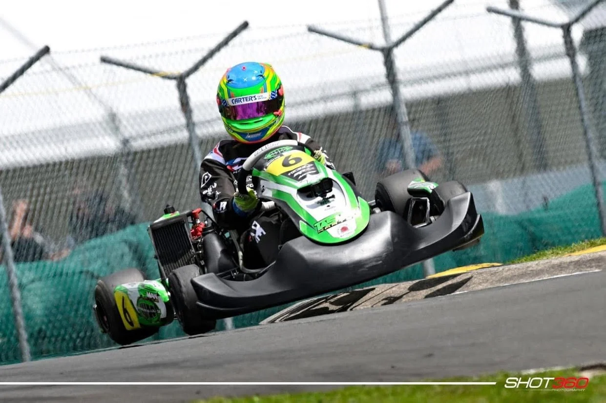 Go-kart racer wearing a colorful helmet and black racing suit driving on a track, with a chain-link fence in the background.