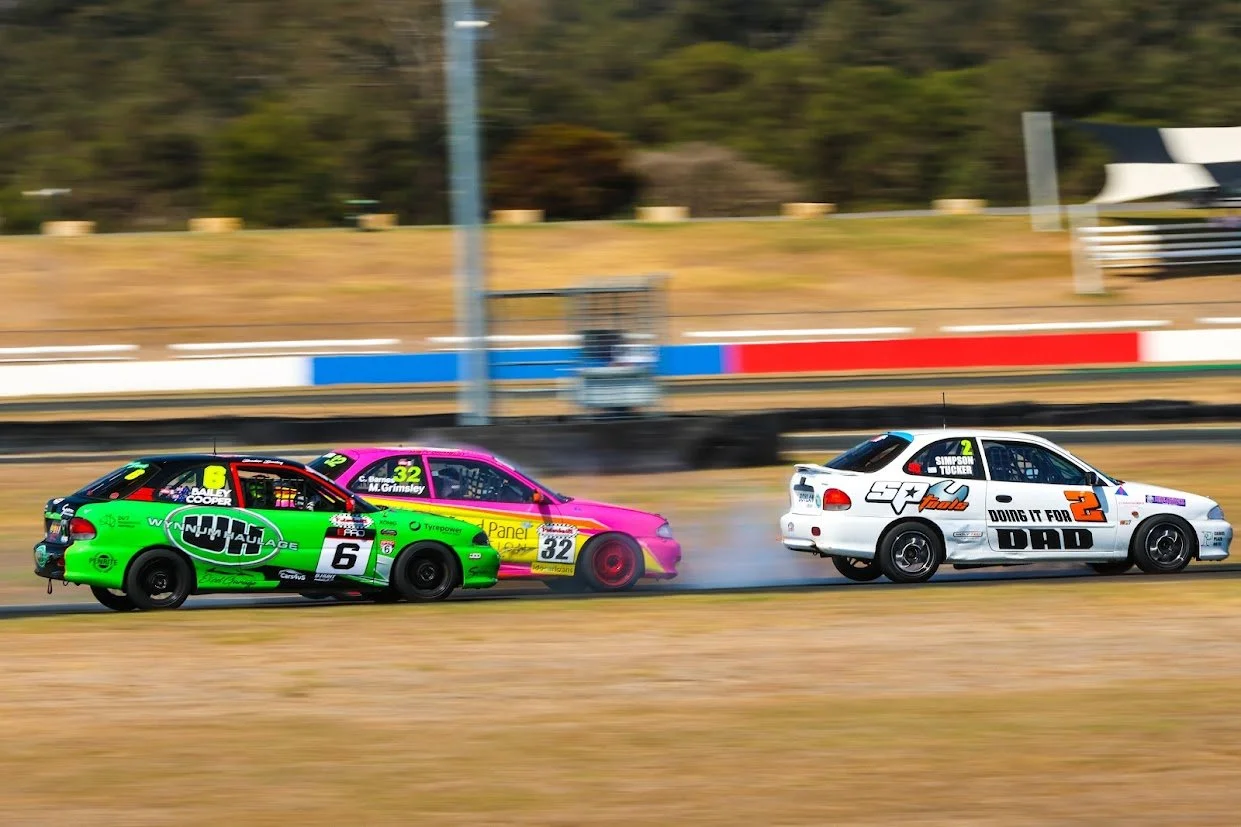 Three race cars on a racetrack, with one car slightly behind the other two. The green car has the number 6, the pink car has the number 32, and the white car has the number 2. Smoke is seen coming from the white car's rear tires.