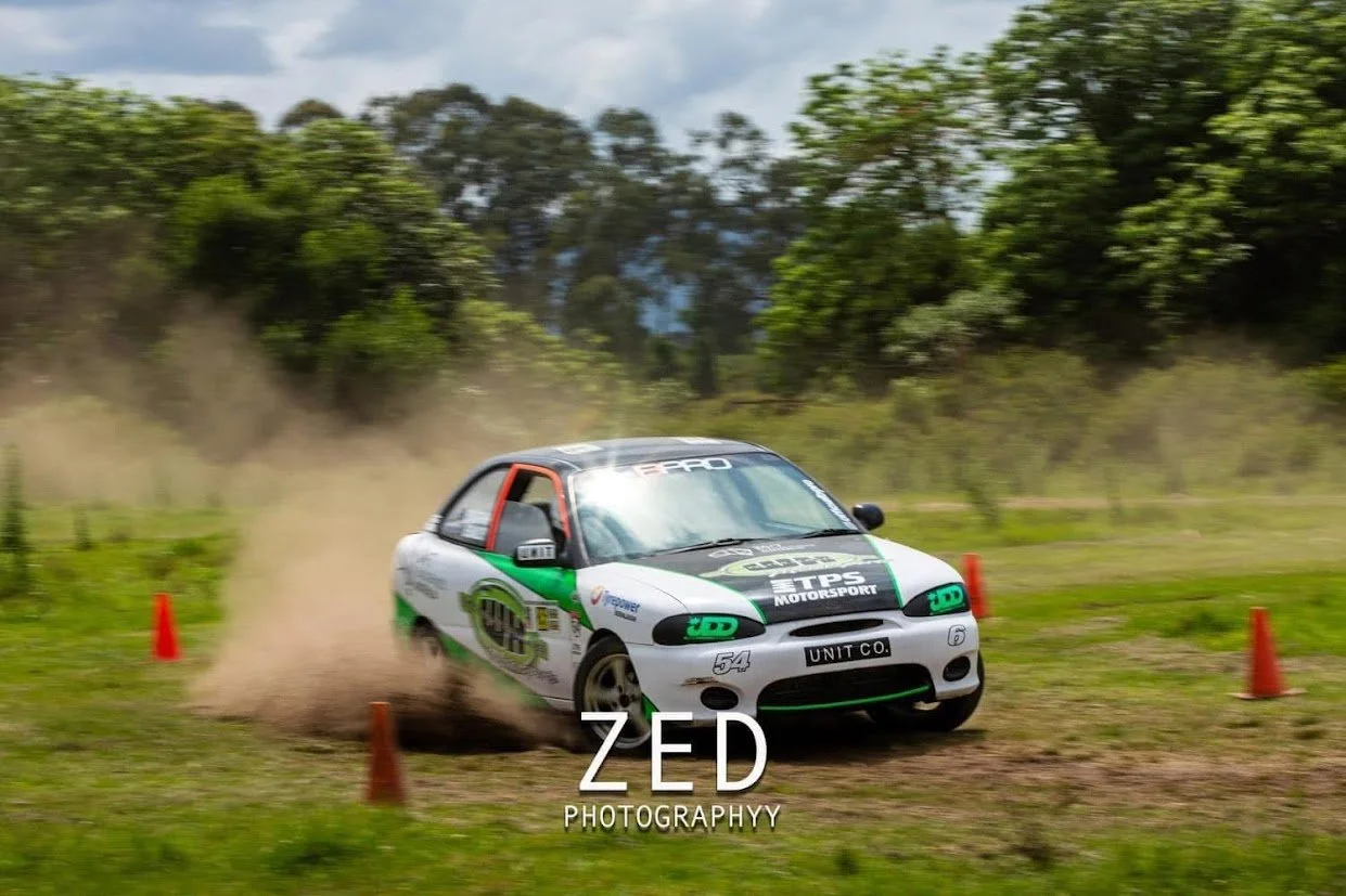 A white and green rally car racing on grass, kicking up dust, around orange cones with trees and cloudy sky in the background.