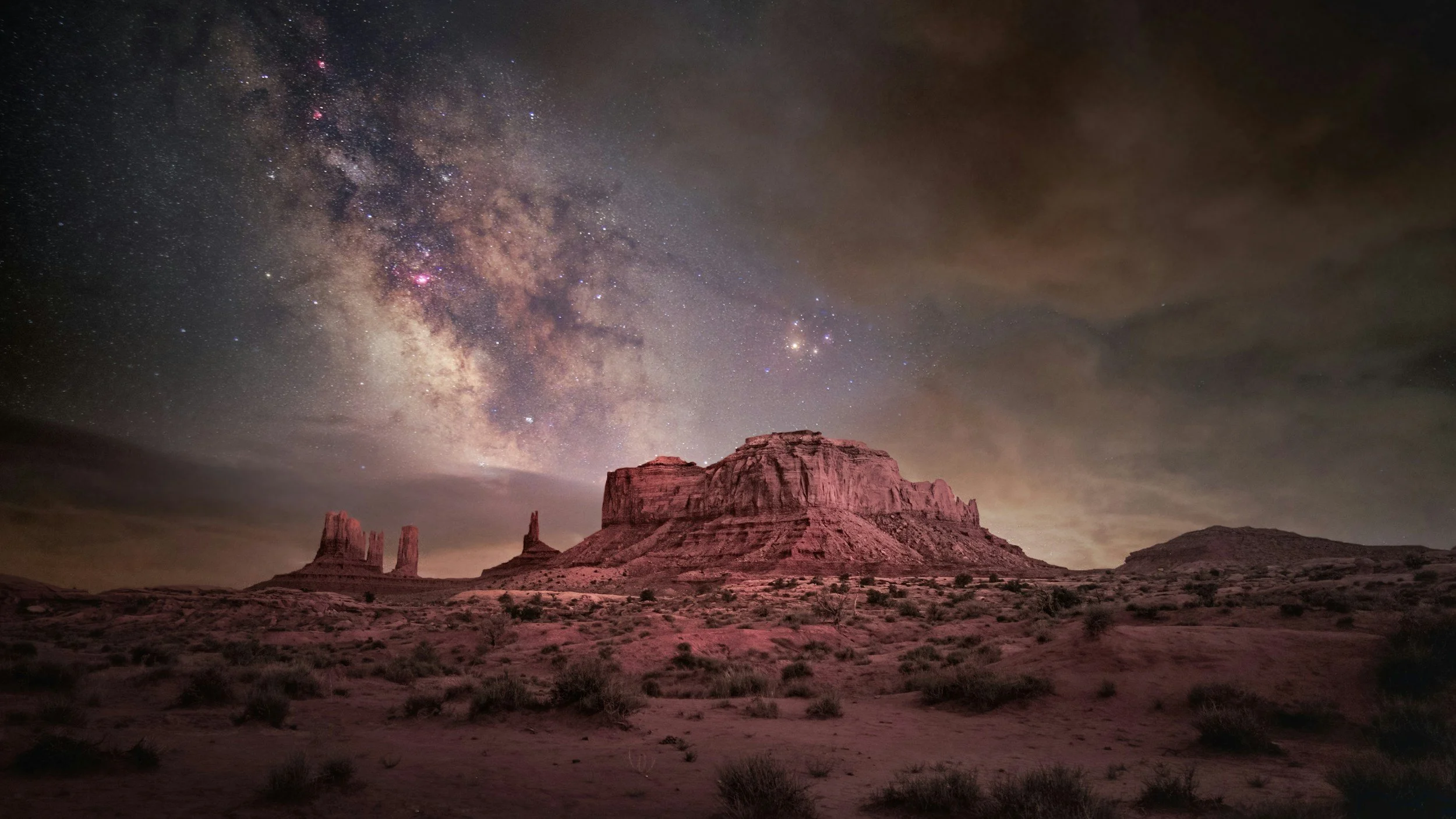 Nighttime view of desert landscape with large rock formations under a starry sky and the Milky Way galaxy visible overhead.