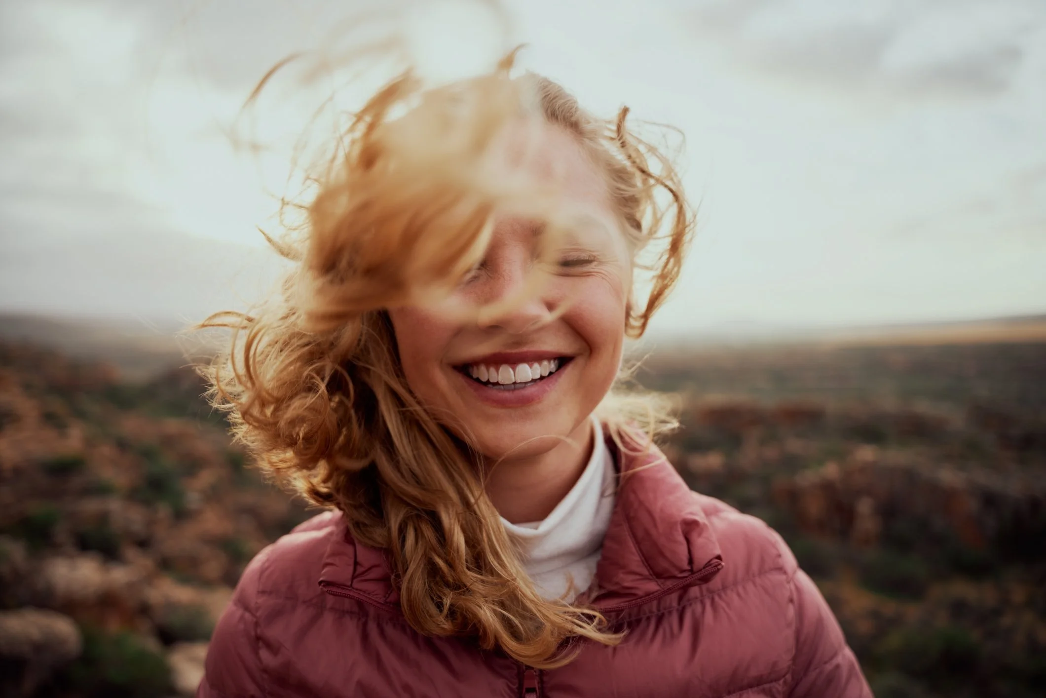 A woman with red hair smiling with her eyes closed, wearing a pink jacket, outdoors in a rocky landscape with a cloudy sky.