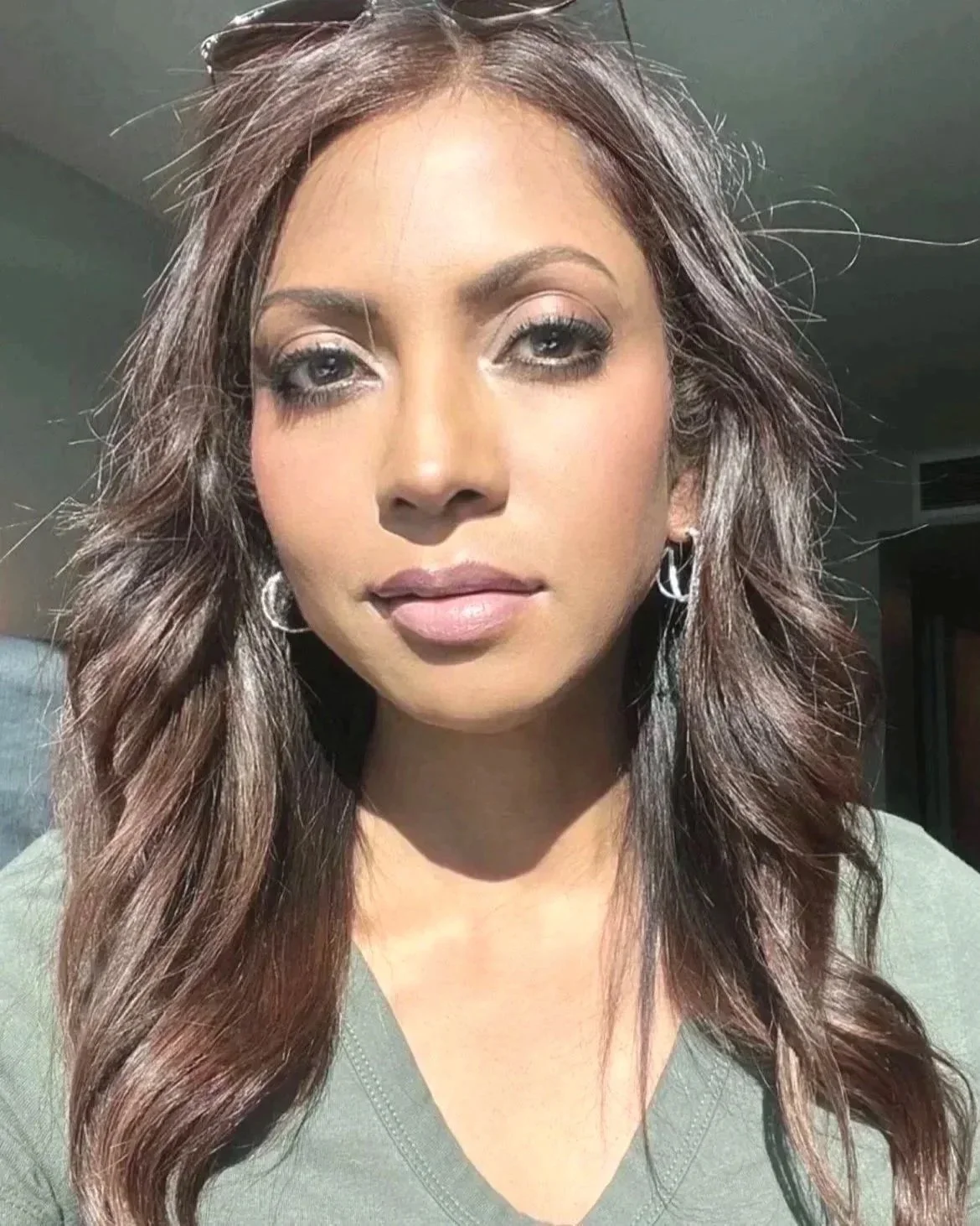 Close-up of a woman with wavy brown hair, wearing earrings, and a green top, standing in sunlight indoors.