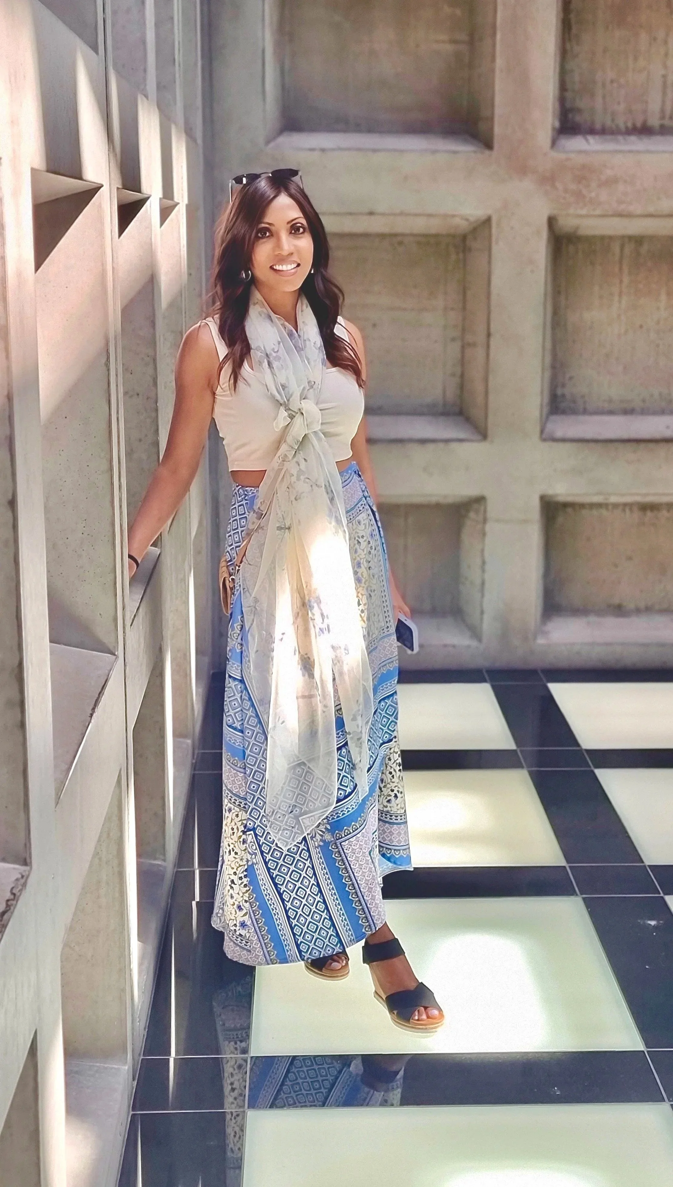A woman standing against a wall with geometric patterns, wearing a sleeveless top, patterned long skirt, and sandals, holding a phone.
