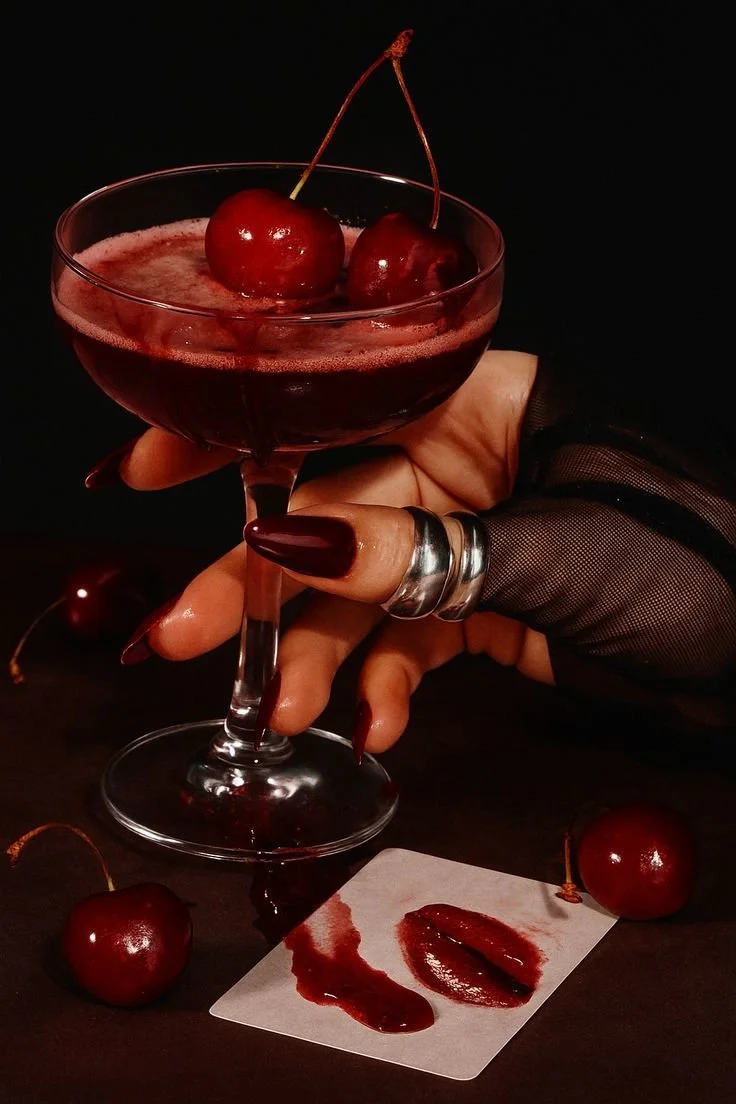A hand with dark red painted nails, wearing rings and a black net glove, holding a cherry cocktail glass with a dark red drink topped with cherries. Several cherries and cherry juice are on a black surface with a white piece of paper underneath, all against a dark background.