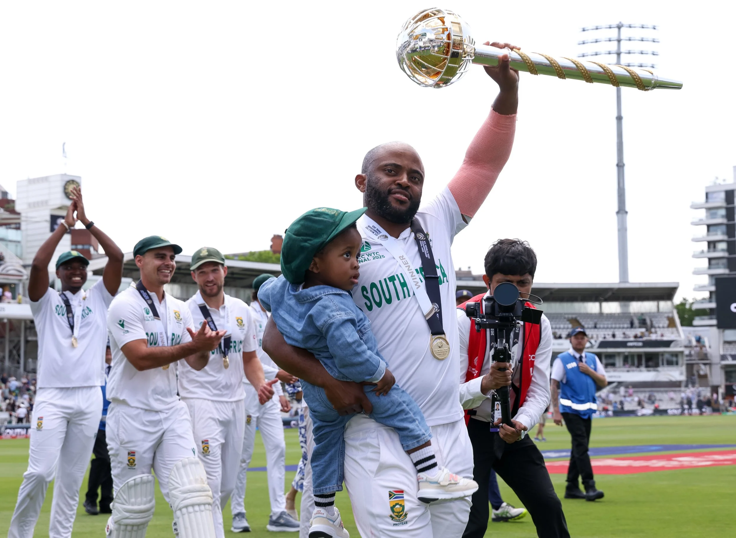 South African cricket team celebrating on the field after winning a match, with one player holding a trophy and a child in his arms.