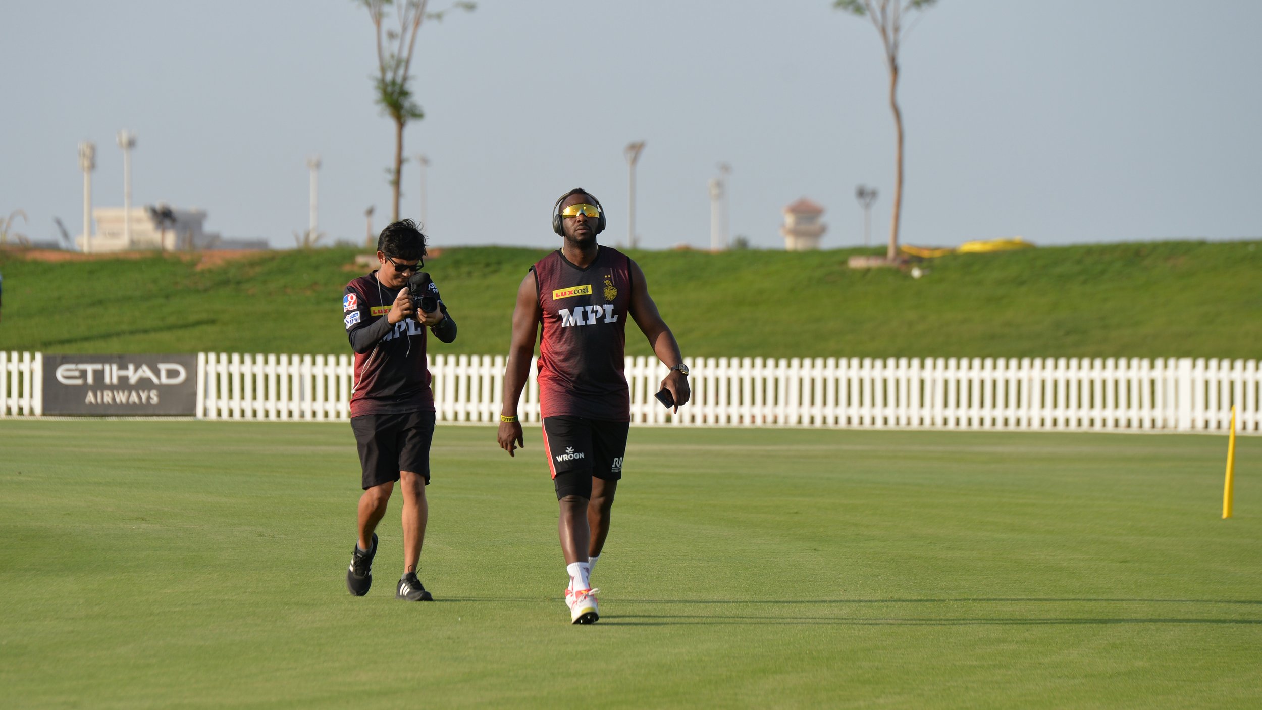 Cricket players walking on the field during a practice session with a fence and some trees in the background.