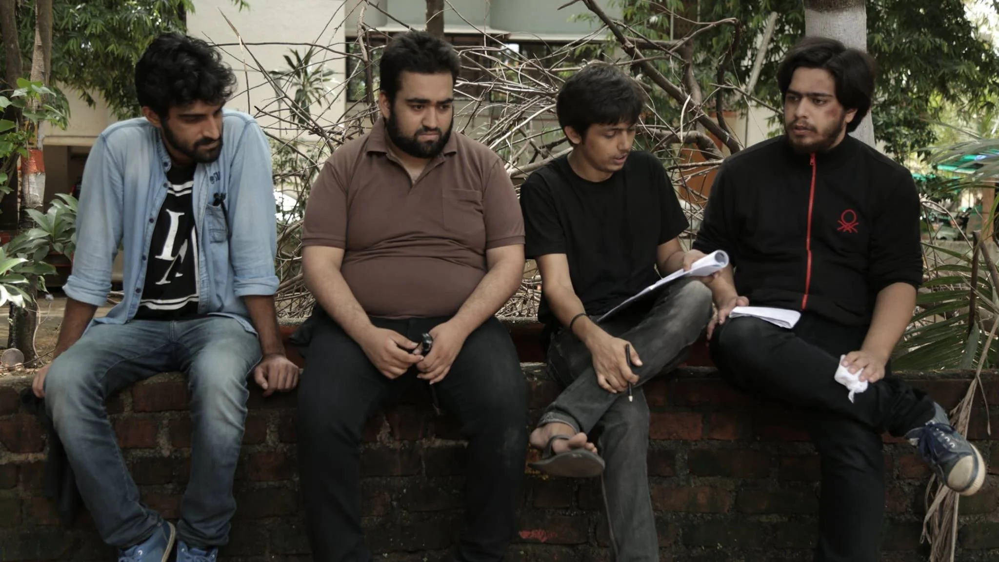 Four young men sitting on a brick wall outdoors, engaged in conversation, with trees and plants in the background.