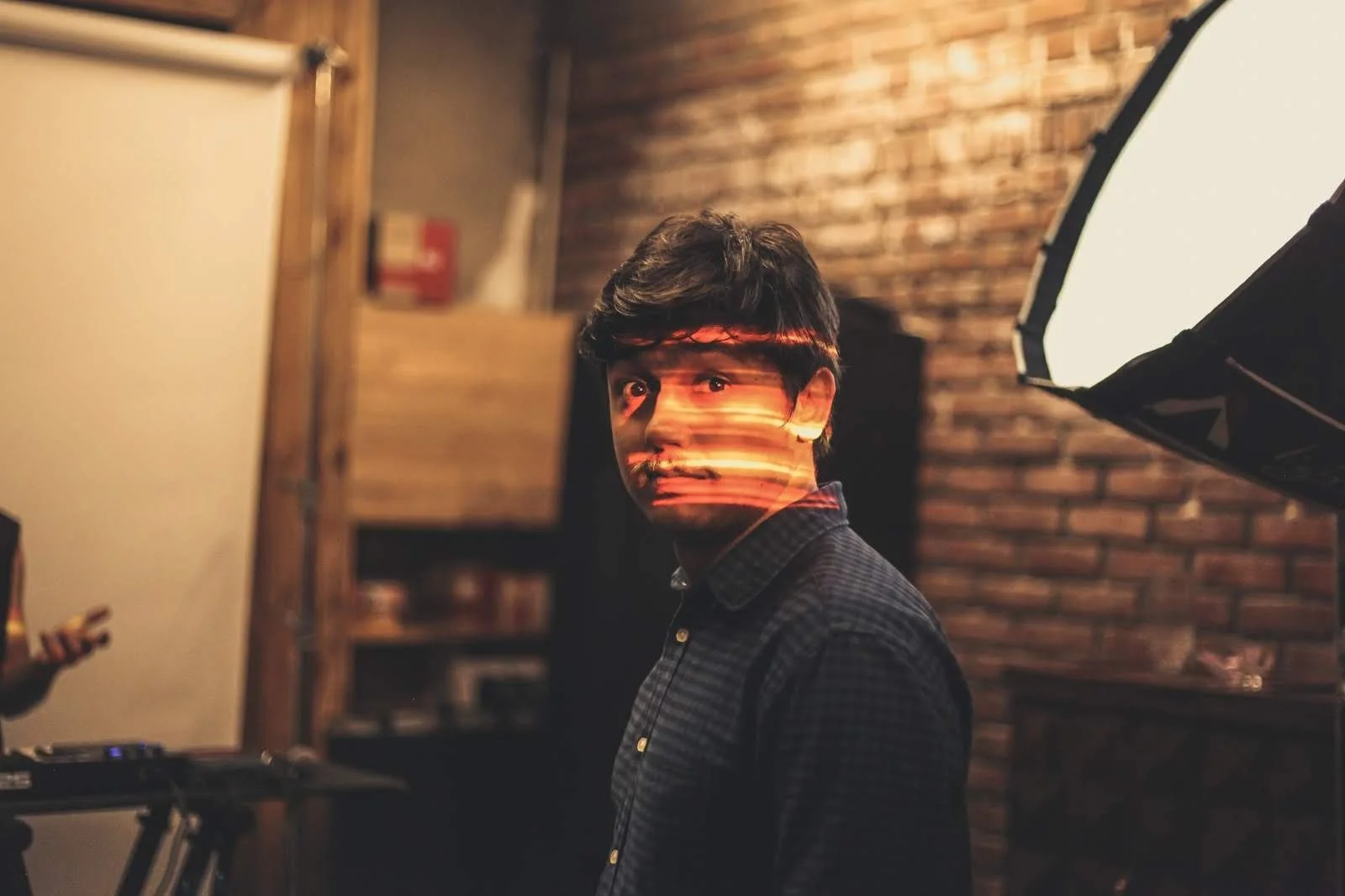 A man standing in a dimly lit room with a brick wall background, illuminated by a light with horizontal streaks reflecting on his face, and a professional lighting setup to the right.