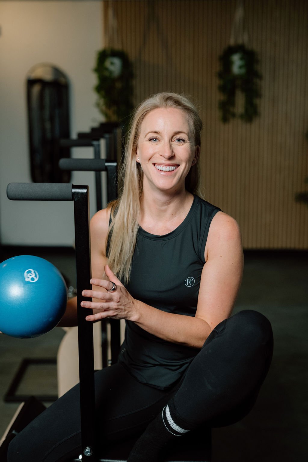 A woman with blonde hair smiling while sitting on the floor in a gym, holding a workout equipment with a blue exercise ball nearby.