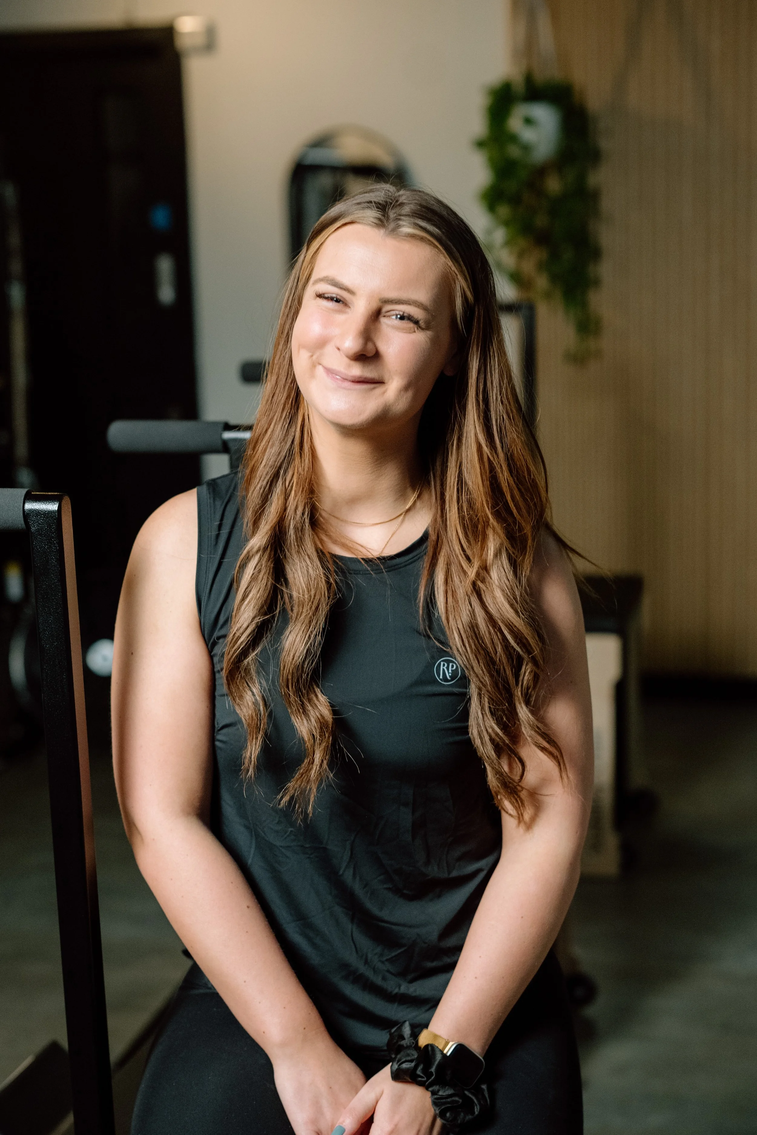 Young woman with long wavy brown hair smiling and winking, sitting in a fitness studio with exercise equipment in the background.