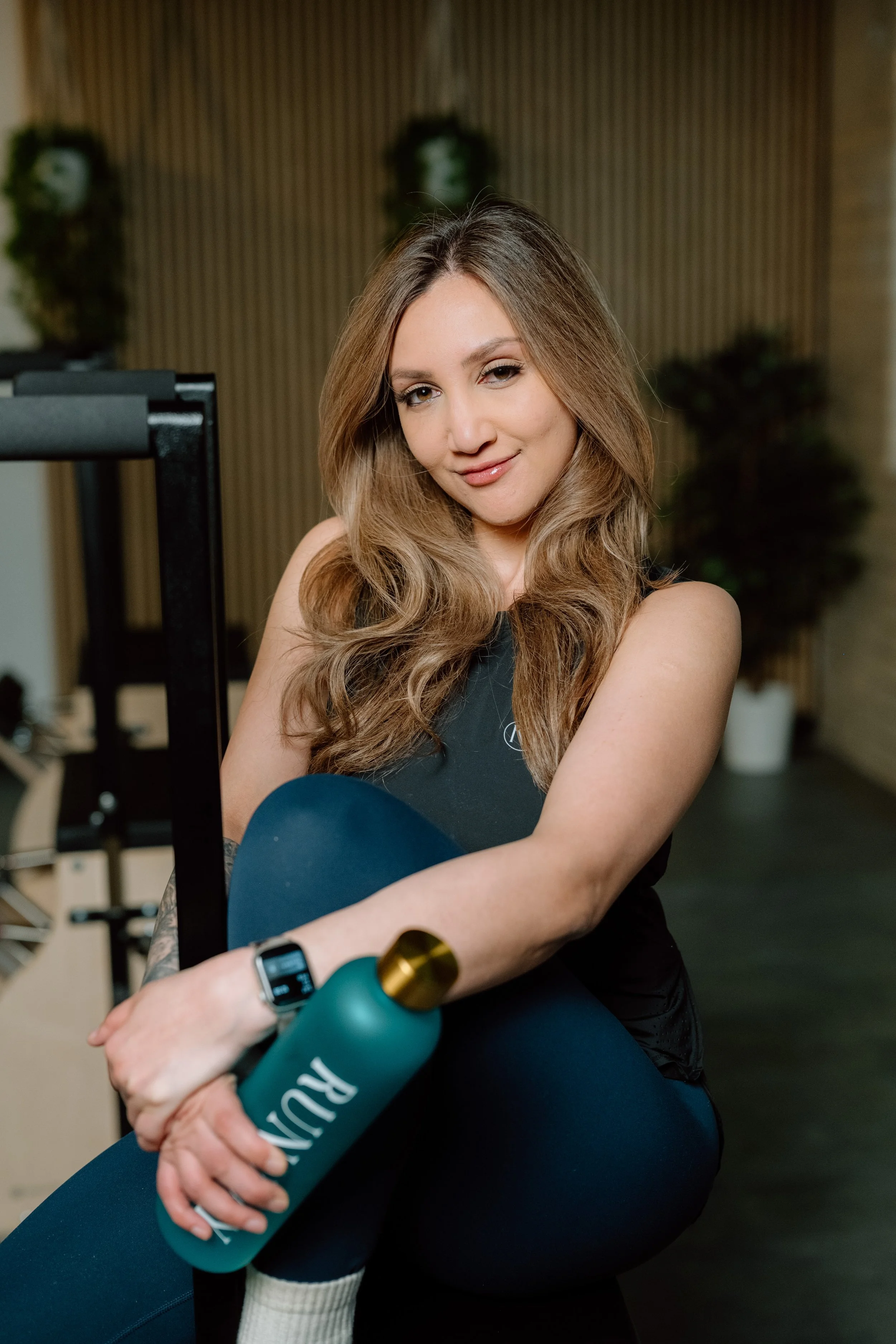 A young woman with long wavy brown hair, wearing a black sleeveless athletic top, holding a blue water bottle, sitting in a gym or fitness studio with workout equipment and plants in the background.