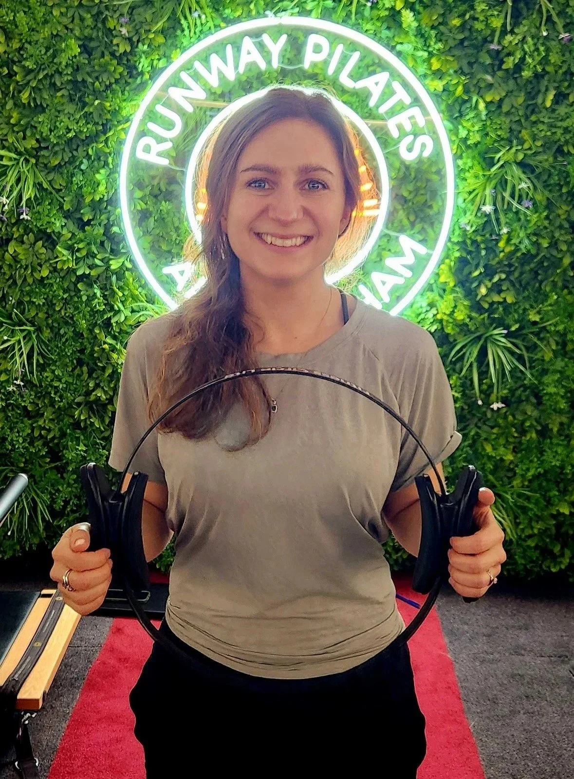 A woman smiling and holding headphones in front of a green wall with a neon sign that reads 'Runway Pilates Miami'.
