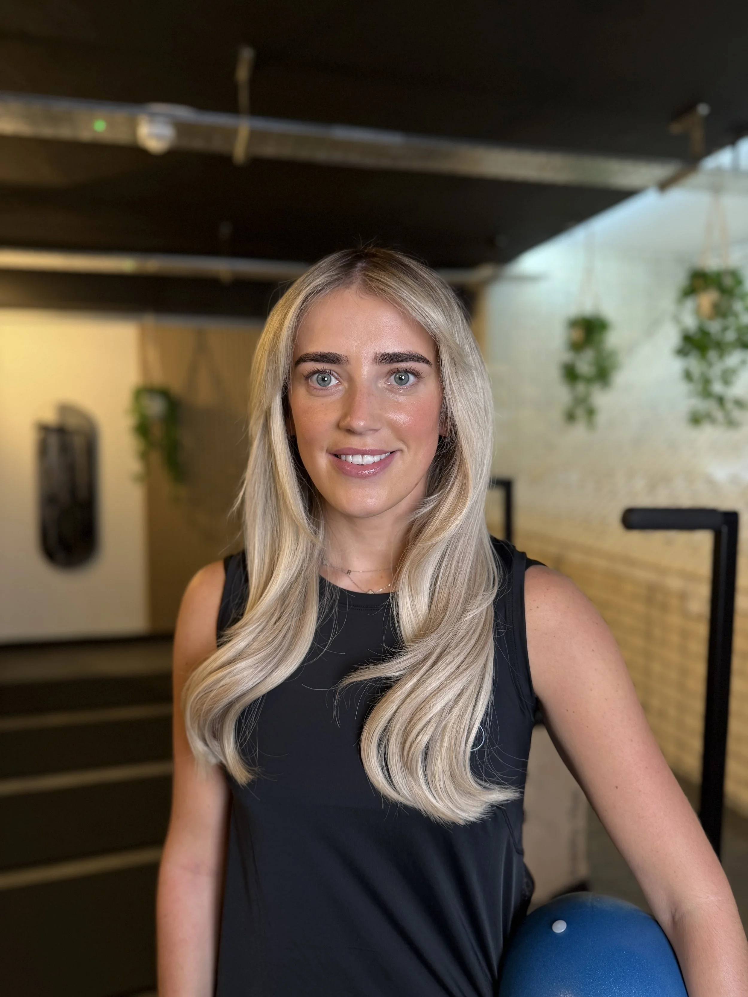 A woman with long blond hair and blue eyes smiling at the camera in an indoor setting with plants hanging in the background.