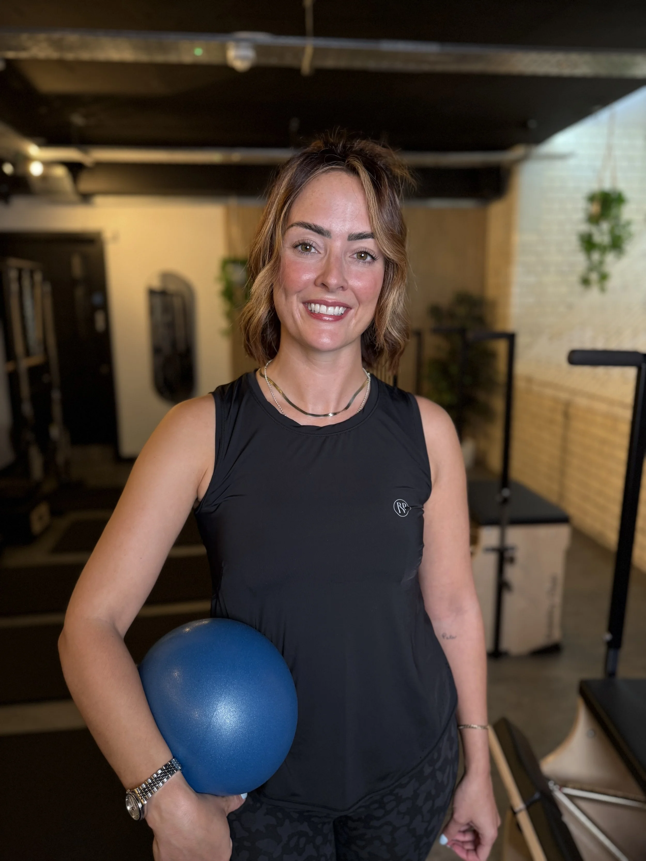 A woman standing in a gym, holding a blue exercise ball under her arm, smiling at the camera, wearing a black athletic tank top and patterned black workout leggings.