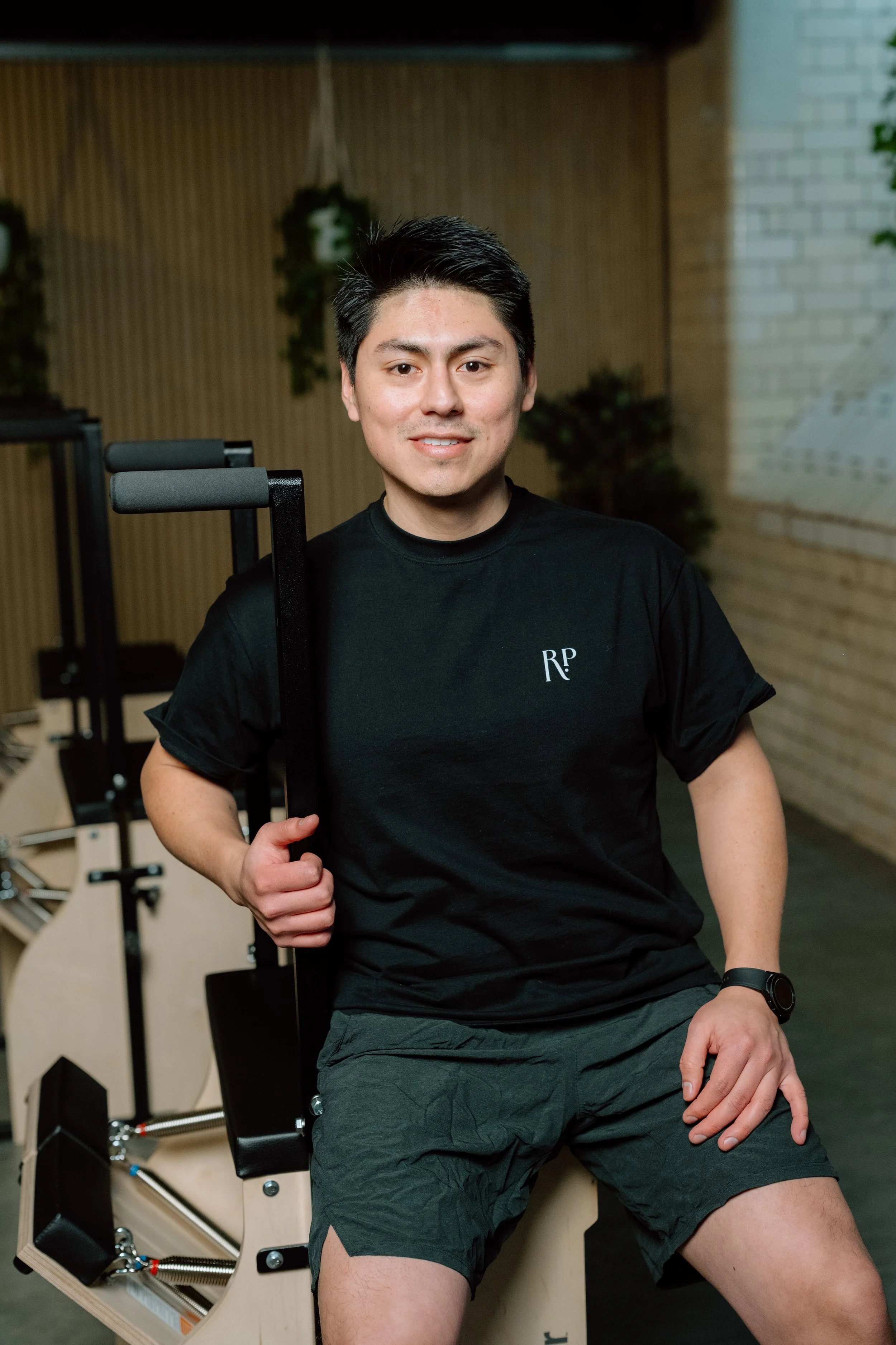Young man in black t-shirt and shorts sitting on a Pilates reformer machine in a gym.