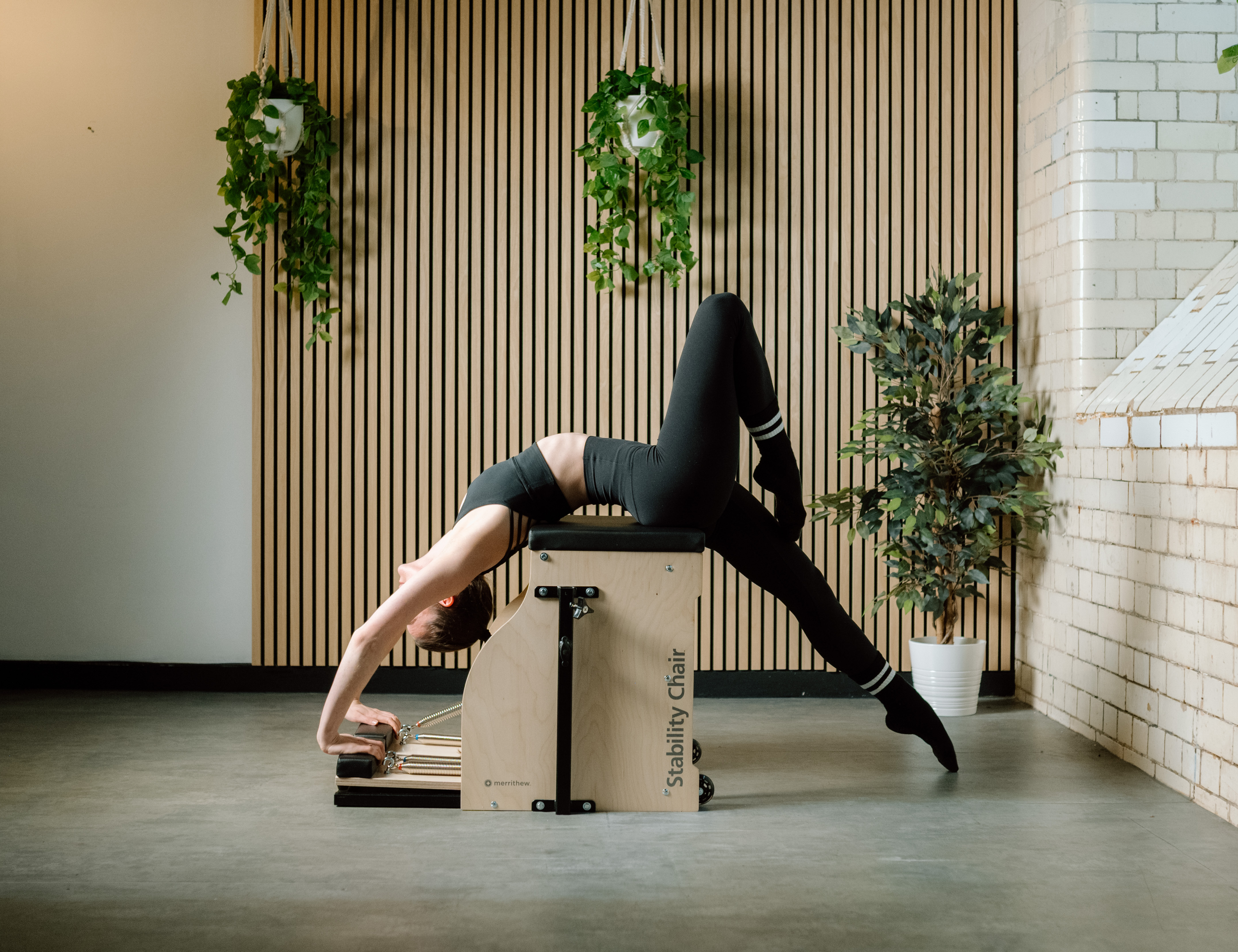 A woman practicing yoga in a studio, performing a bridge pose on a wooden stability chair with plants and wooden wall decor in the background.