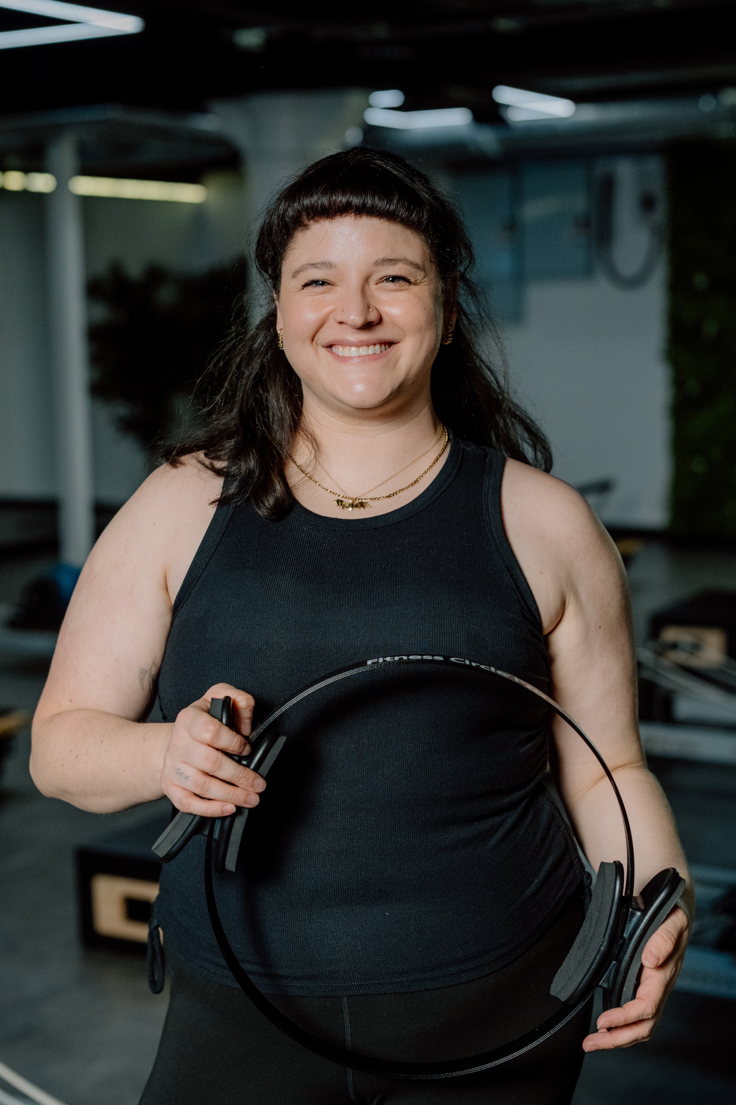 Woman smiling in a gym, holding a black fitness headband, wearing black sleeveless workout attire and layered necklaces.