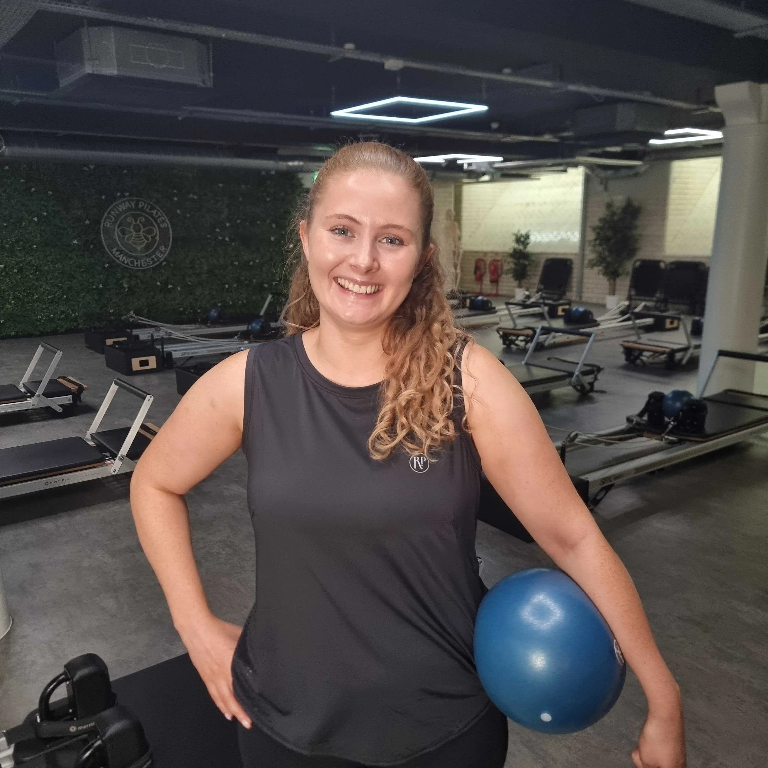 A smiling woman with curly hair holding a blue exercise ball in a gym with workout equipment in the background.