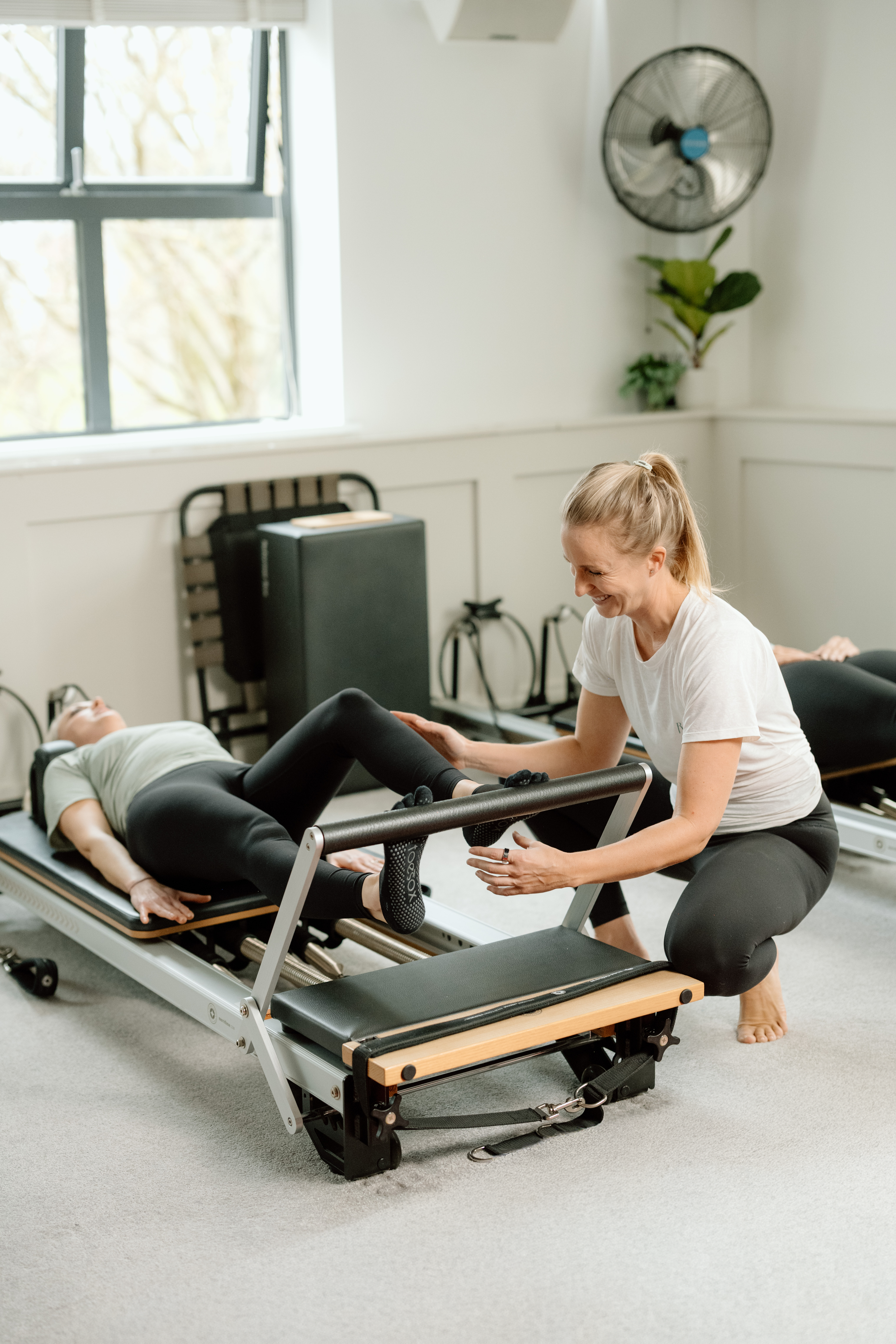 A woman is assisting a person with a Pilates exercise on a reformer machine in a bright room with a window, fan, and plants.