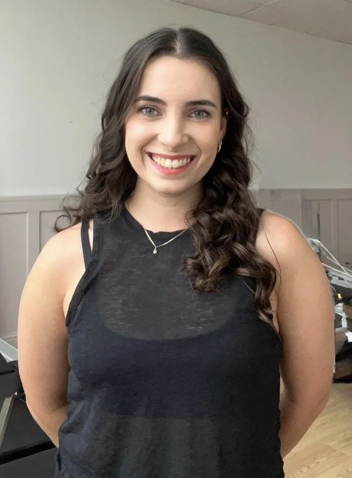 Young woman with long wavy brown hair smiling and winking, sitting in a fitness studio with exercise equipment in the background.