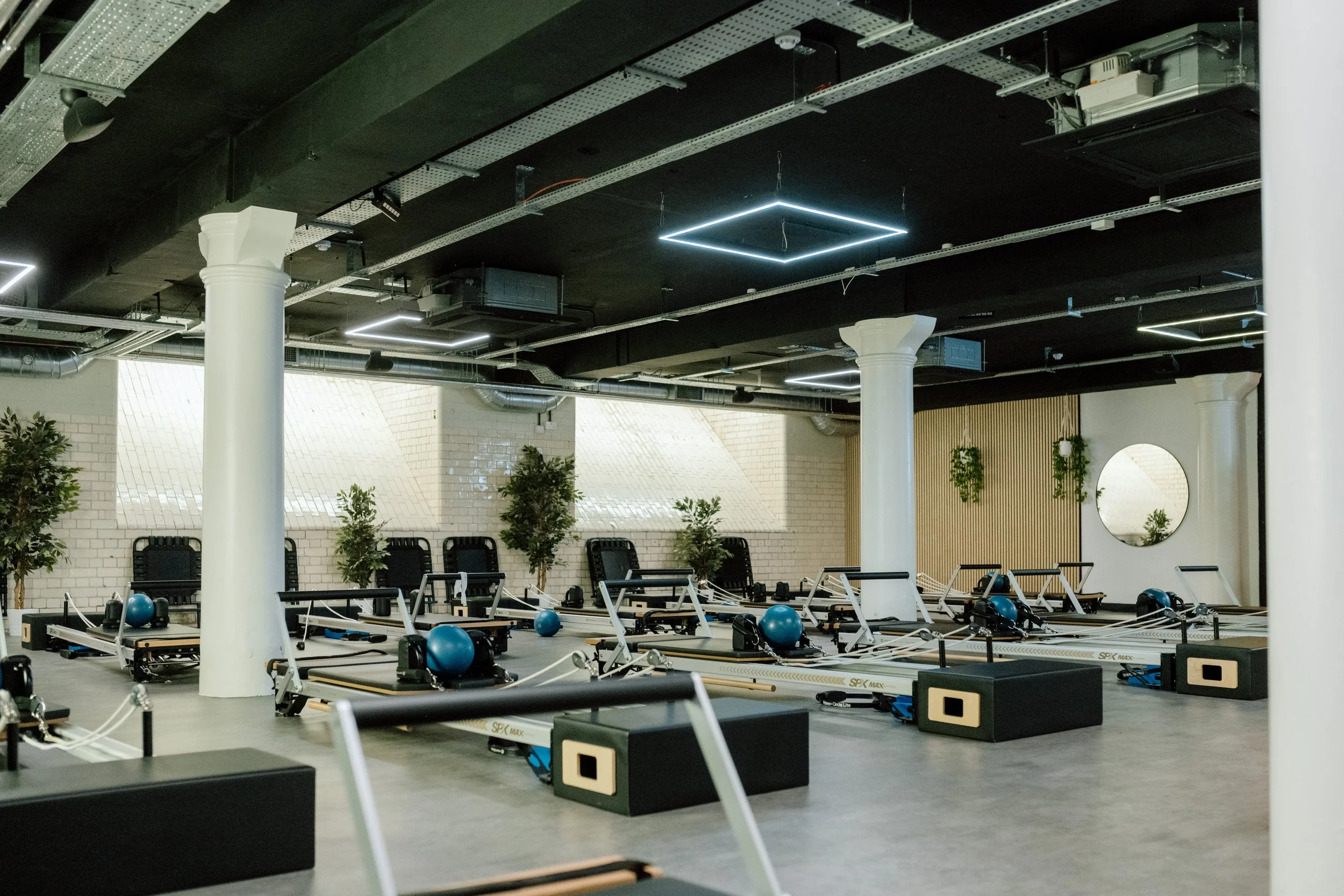 Indoor Pilates or fitness studio with reformer machines, black and white decor, potted plants, and modern lighting fixtures.