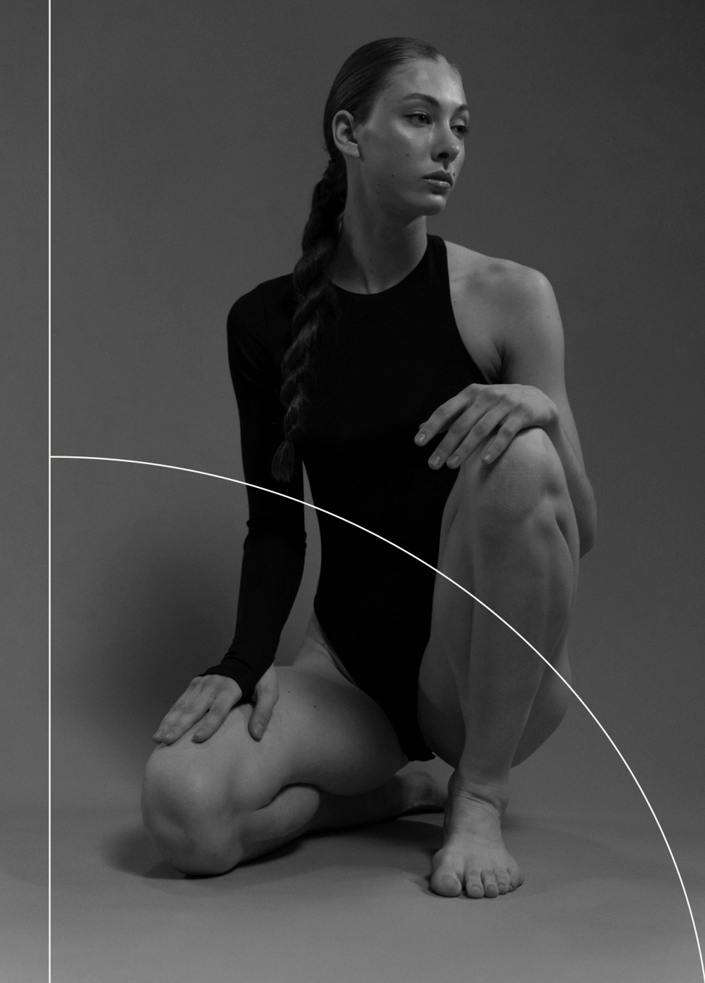 A woman with long dark hair in a braid, wearing a black leotard, seated on the floor with one knee raised and her hand resting on her knee.