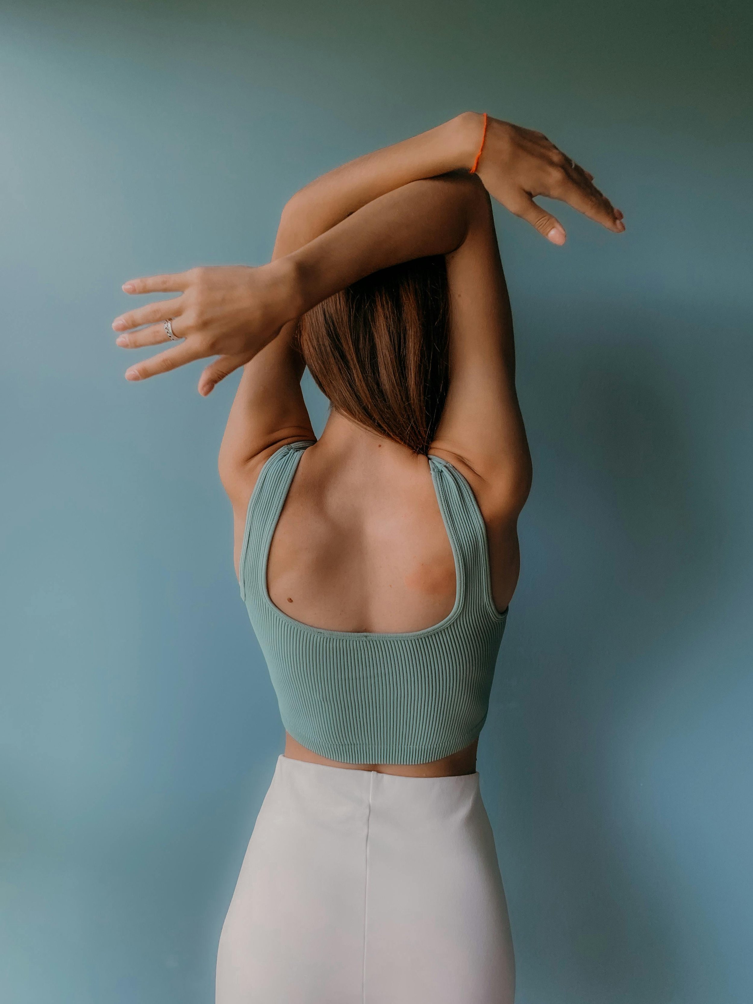 Woman with brown hair wearing a ribbed green tank top and a white skirt, stretching her arms over her head against a plain blue background.