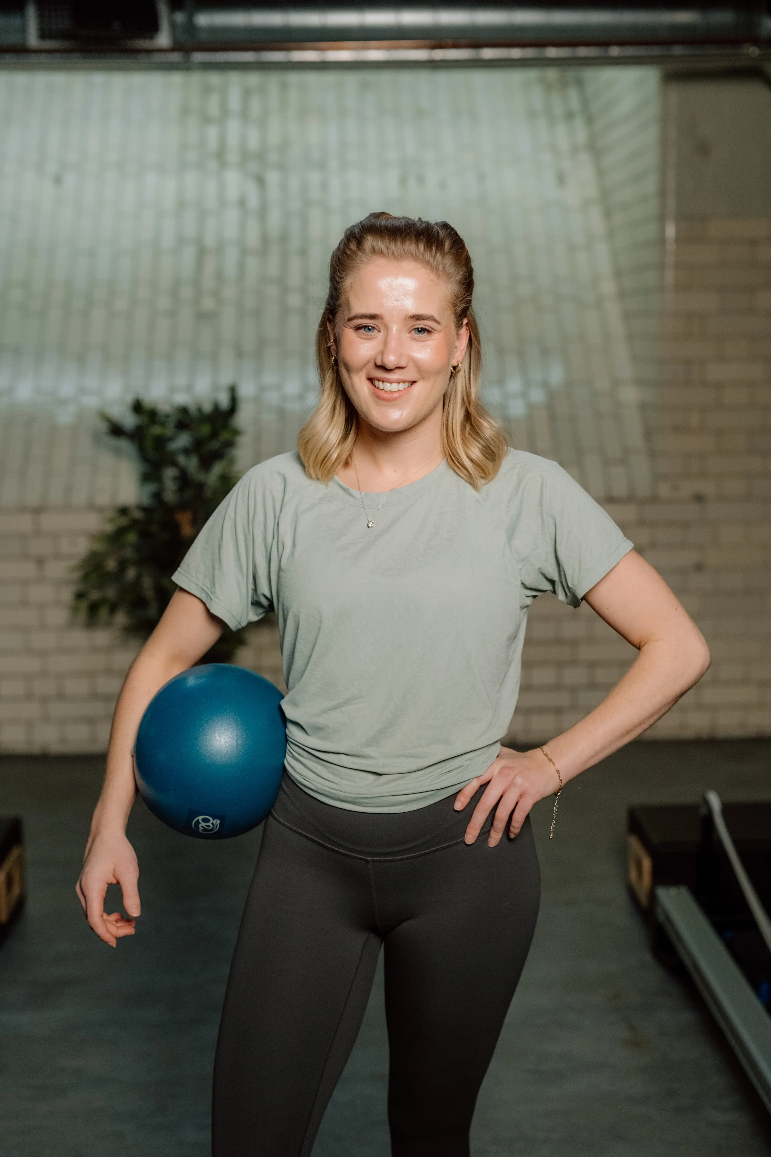 A young woman in workout attire standing in a gym, smiling, holding a blue exercise ball, with gym equipment in the background.