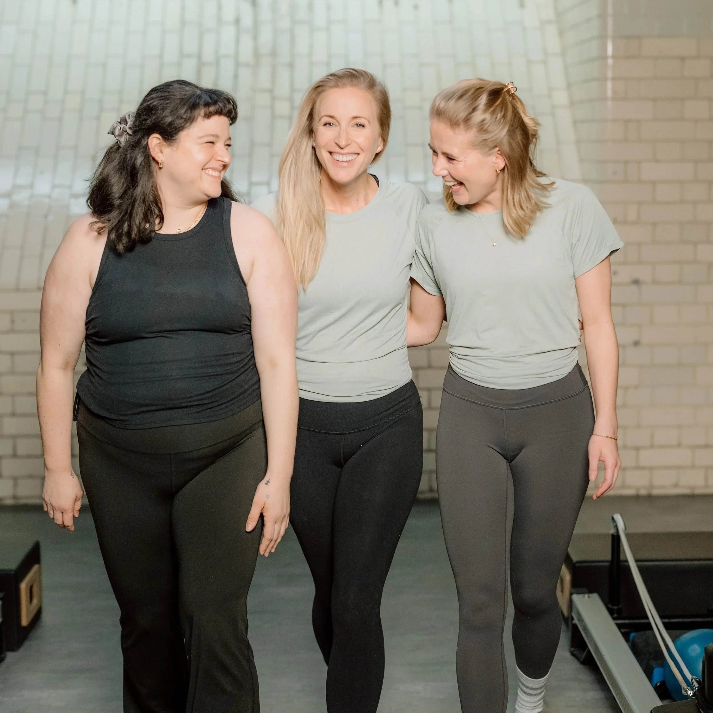 Three women smiling and walking together in a gym with a brick wall background.