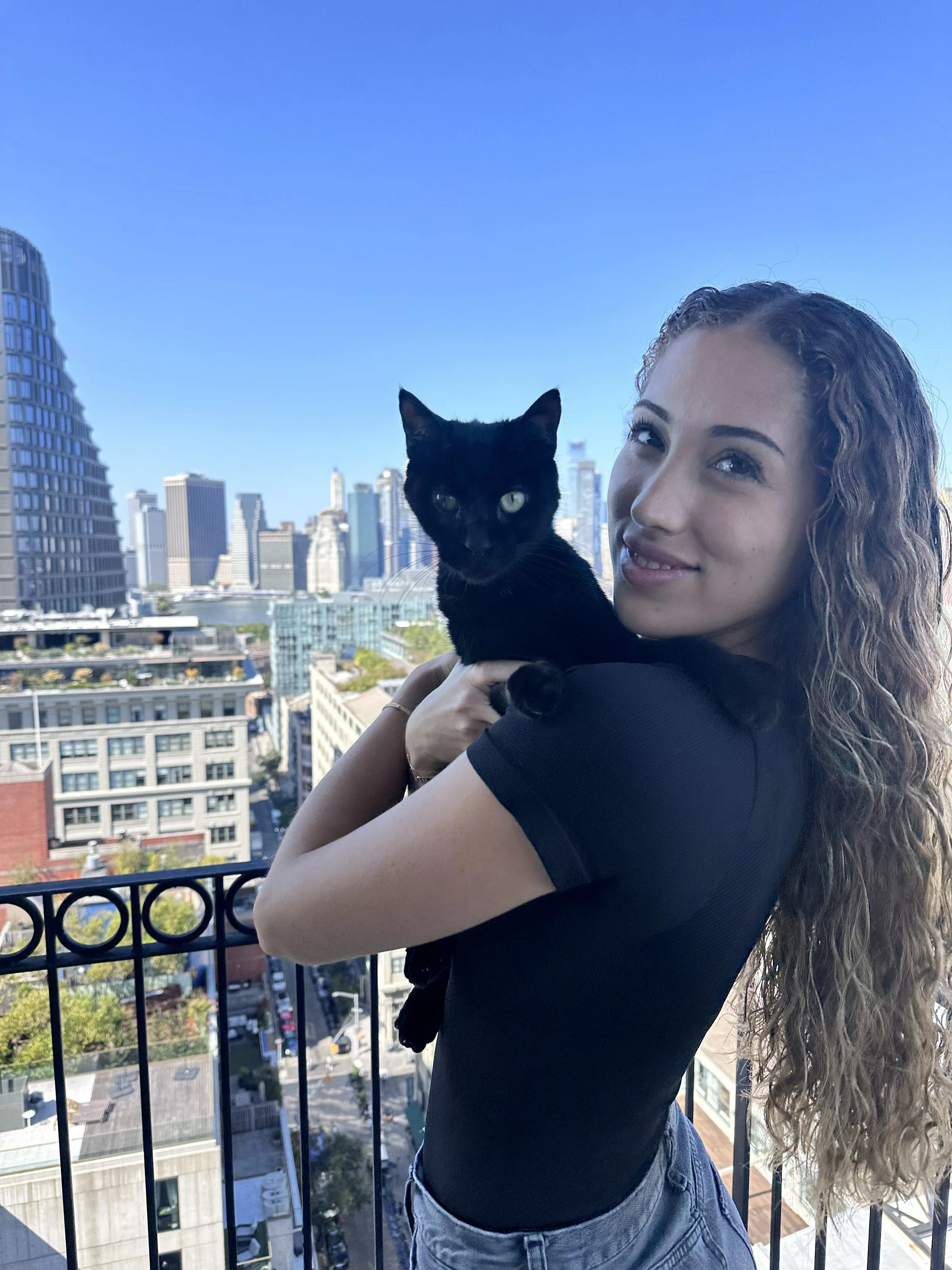 A young woman with long curly hair holding a black cat on a balcony with a city skyline in the background.
